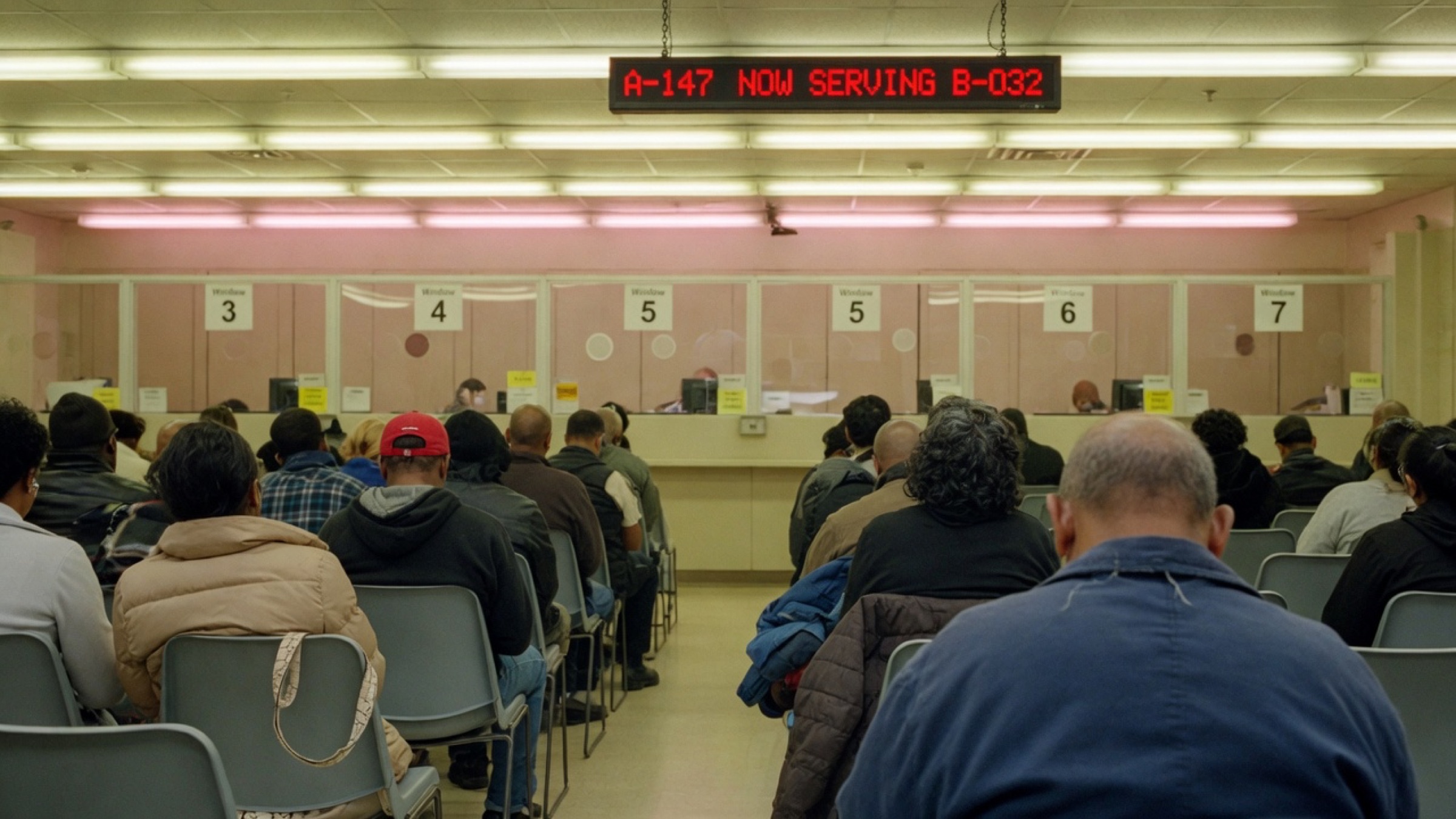 People sitting in a DMV waiting room, seen from behind, facing numbered windows and a Now Serving display.